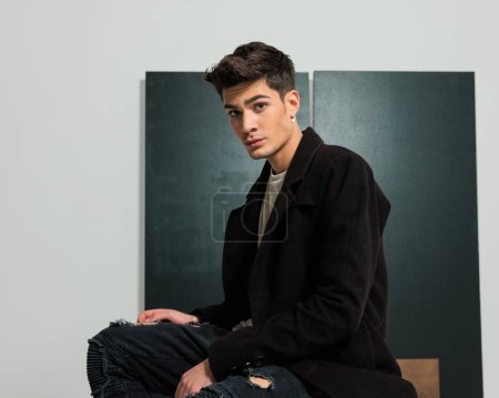 Foto de Vista lateral de hombre joven de moda casual en abrigo negro con jeans sentados en el estudio mientras mira hacia adelante y posando sobre fondo gris - Imagen libre de derechos