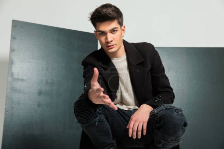 Photo for Sexy casual man in black coat crouching, looking forward and shaking hand, posing in a fashion way in front of grey background - Royalty Free Image