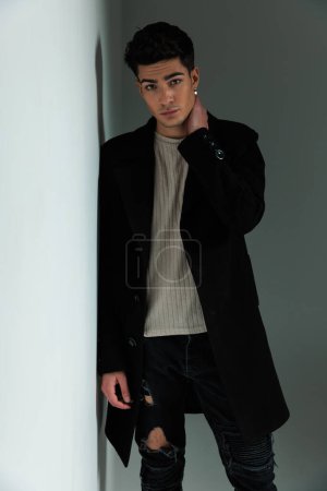 Foto de Hombre de moda fresco en abrigo negro cogido de la mano en el cuello mientras se apoya en una pared y misteriosamente posando en el estudio - Imagen libre de derechos
