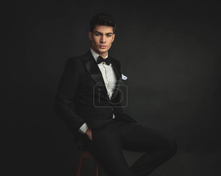 Photo for Close up of relaxed gentleman wearing black suit sitting on chair with hands in pockets - Royalty Free Image