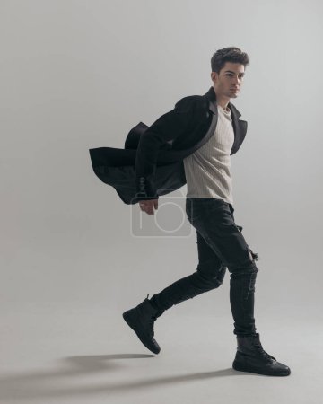 Téléchargez les photos : Attrayant jeune homme marchant sur le côté avec un manteau noir flottant dans l'air isolé sur fond blanc - en image libre de droit