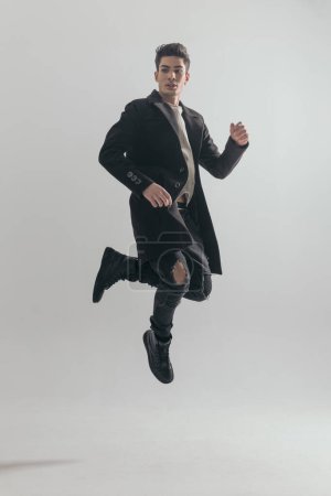 Photo for Young guy wearing black overcoat leaping up in the air on light grey background - Royalty Free Image