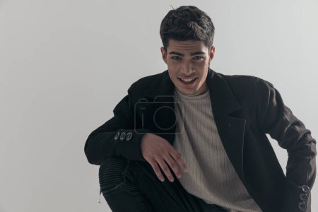 Photo for Closeup of happy young man in black coat crouching down on grey background - Royalty Free Image