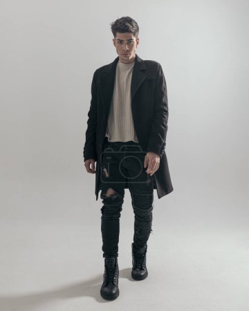 Foto de Joven confiado y sexy con abrigo negro y jeans avanzando sobre fondo gris - Imagen libre de derechos