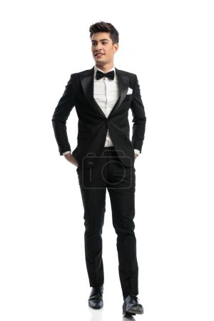 Photo for Full length picture of happy elegant groom in black tuxedo looking to side and smiling while holding hands in pockets on white background - Royalty Free Image