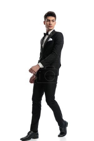Téléchargez les photos : Bel homme élégant en smoking noir marchant et regardant derrière tout en posant avec les bras dans la pose de mode sur fond blanc - en image libre de droit