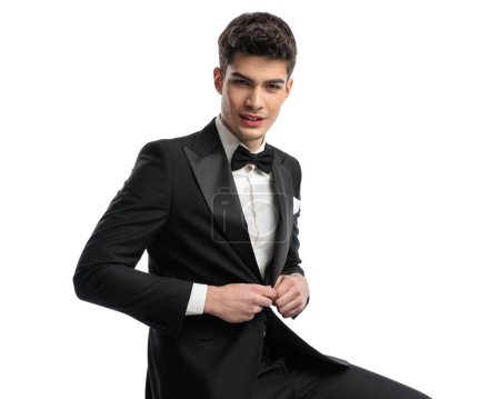 Photo for Portrait of happy young man smiling while unbuttoning black tuxedo and sitting in front of white background - Royalty Free Image