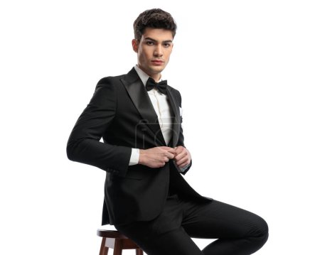 Photo for Portrait of seated elegant groom arranging and buttoning black tuxedo in front of white background in studio - Royalty Free Image