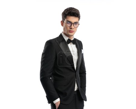 Photo for Portrait of sexy young groom with glasses in black tuxedo standing with hands in pockets and posing on white background - Royalty Free Image
