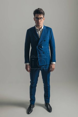 Téléchargez les photos : Image complète du corps de l'homme élégant en costume bleu triple poitrine debout et posant sur fond gris - en image libre de droit
