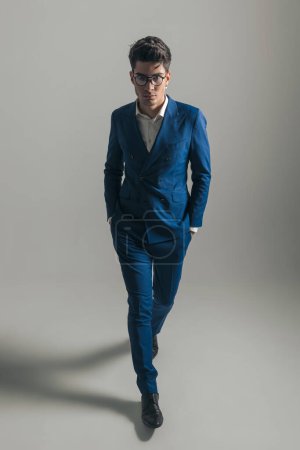 Téléchargez les photos : Image complète du corps de cool homme d'affaires de mode en costume bleu triple poitrine marchant avec les mains dans les poches devant fond gris - en image libre de droit