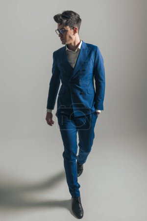 Téléchargez les photos : Image complète du corps de l'homme élégant en costume bleu triple poitrine avec des lunettes marchant avec les mains dans les poches et regardant sur le côté sur fond gris - en image libre de droit