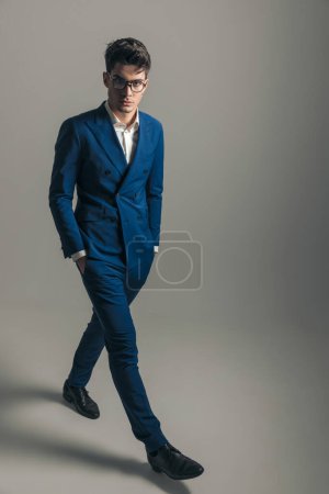 Téléchargez les photos : Cool homme d'affaires de mode en costume bleu triple poitrine marchant avec les mains dans les poches en face de fond gris - en image libre de droit