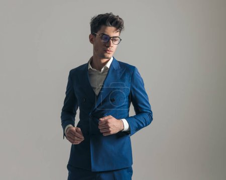 Téléchargez les photos : Portrait d'un bel homme confiant en costume bleu avec des lunettes regardant loin et posant devant un fond gris - en image libre de droit