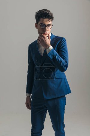 Téléchargez les photos : Réfléchi jeune homme d'affaires en costume bleu triple poitrine toucher le menton, penser et détourner les yeux sur fond gris - en image libre de droit