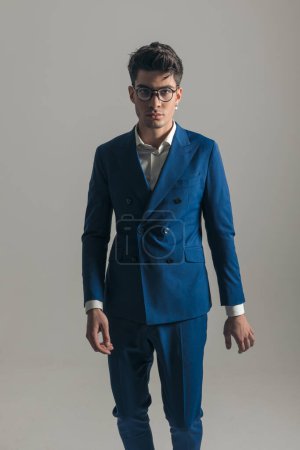 Téléchargez les photos : Portrait d'un homme d'affaires confiant avec des lunettes portant un costume bleu triple poitrine et posant d'une manière cool devant un fond gris - en image libre de droit