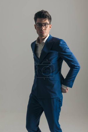 Téléchargez les photos : Cool homme de mode avec des lunettes en costume tenant la main derrière et posant devant fond gris - en image libre de droit