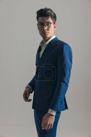 Téléchargez les photos : Vue de côté photo de l'homme d'affaires élégant en costume bleu triple poitrine avec des lunettes debout et posant en face de fond gris - en image libre de droit
