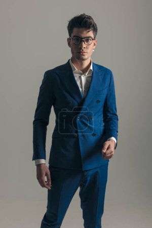 Téléchargez les photos : Bel homme d'affaires élégant en costume bleu avec des lunettes debout et regardant vers l'avant en face de fond gris - en image libre de droit