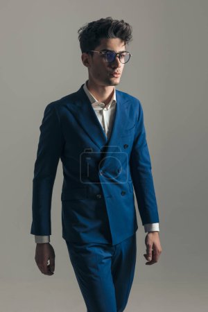 Téléchargez les photos : Sexy jeune homme avec des lunettes en costume bleu triple poitrine marchant et regardant loin sur fond gris - en image libre de droit