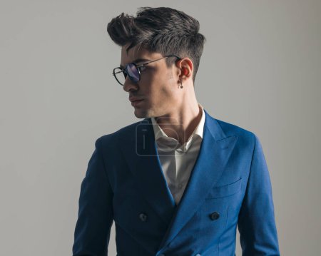 Téléchargez les photos : Portrait d'élégant jeune homme élégant en costume bleu avec des lunettes regardant vers le côté et posant devant fond gris - en image libre de droit