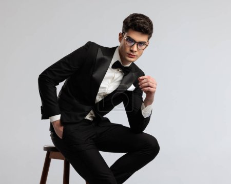 Photo for Close up of sexy groom in black tux sitting on wooden chair and leaning forward on gray background - Royalty Free Image