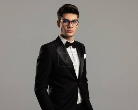 Foto de Retrato de novio guapo con esmoquin negro y gafas de pie sobre fondo gris con manos en bolsillos - Imagen libre de derechos