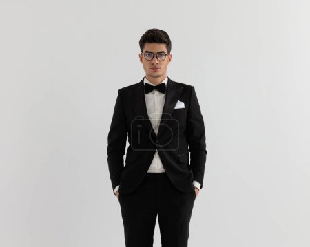 Photo for Closeup of relaxed young man in black suit standing with hands in pockets while wearing glasses on gray background - Royalty Free Image