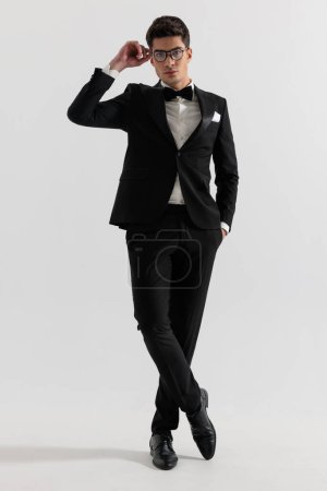 Photo for Relaxed fashion man fixing his glasses while standing with folded feet on gray background - Royalty Free Image