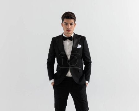 Photo for Closeup of sexy fashion man wearing black suit holding pockets while standing on gray background - Royalty Free Image