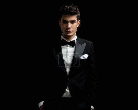 Photo for Closeup of young man wearing tuxedo and bowtie standing on black background with hands in pockets - Royalty Free Image