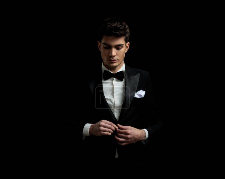 Photo for Portrait of handsome young man buttoning his black tux and looking down on black background - Royalty Free Image
