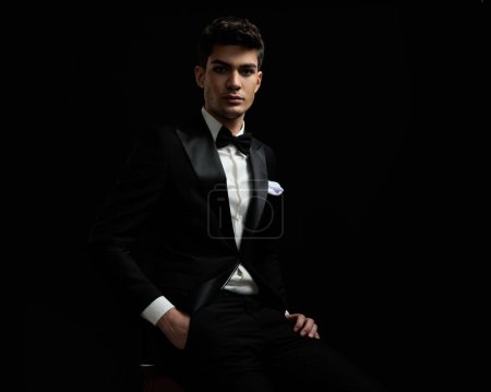 Photo for Portrait of young businessman wearing black suit and bow tie sitting with hand in pocket on dark background - Royalty Free Image