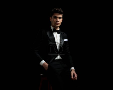 Photo for Young fashion man wearing black suit and bowtie resting with hand in pocket on chair on black background - Royalty Free Image