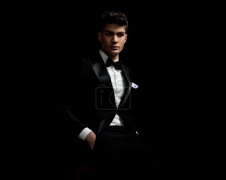 Photo for Attractive groom in black tuxedo looking to side while sitting on a chair on black background - Royalty Free Image