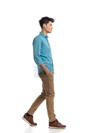 Photo for Side view of relaxed handsome man wearing blue shirt stepping on white background with hands in pockets - Royalty Free Image
