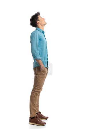 Foto de Vista lateral de un hombre casual joven relajado mirando hacia arriba mientras está de pie en línea sobre un fondo blanco con las manos en los bolsillos - Imagen libre de derechos