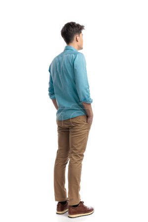 rear view of relaxed casual man wearing blue shirt standing on white background with hands in pockets