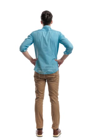 Photo for Back view of young casual man holding hips and looking furious while standing on white background - Royalty Free Image
