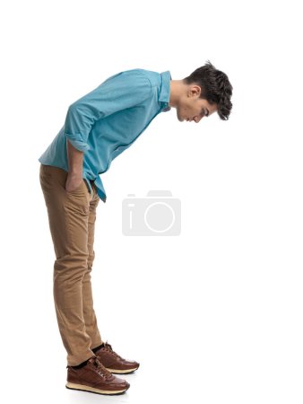 Foto de Vista lateral del relajado hombre casual inclinándose hacia adelante y mirando hacia abajo mientras está de pie con las manos en bolsillos sobre fondo blanco - Imagen libre de derechos