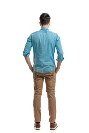 vista trasera del relajado hombre casual con camisa azul y pantalones vaqueros sosteniendo bolsillos mientras está de pie sobre fondo blanco