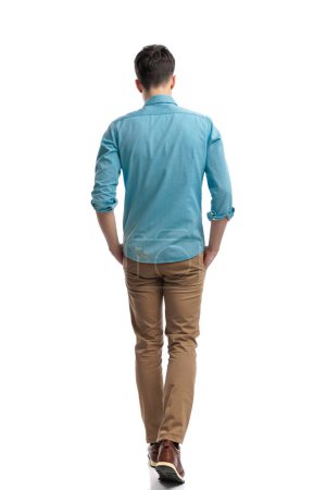 Téléchargez les photos : Vue arrière de l'homme décontracté détendu en chemise bleue marchant vers l'avant sur fond blanc - en image libre de droit