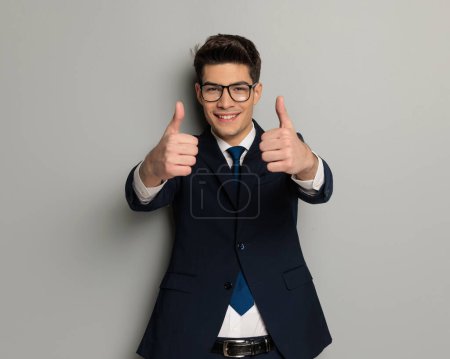 Photo for Closeup of handsome fashion man making thumbs up sign while standing on grey background in black suit - Royalty Free Image