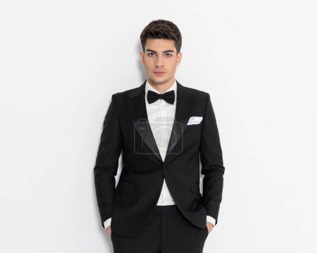 Photo for Elegant young businessman in black tuxedo holding hands in pockets and looking forward in front of grey background - Royalty Free Image
