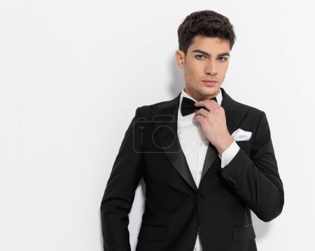 Téléchargez les photos : Portrait de jeune homme d'affaires sexy en smoking noir ajustant noeud papillon noir et regardant vers l'avant devant fond gris - en image libre de droit