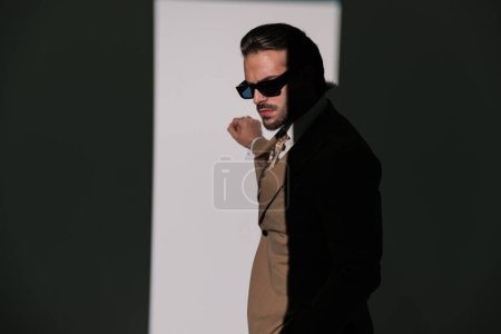 Foto de Vista lateral de elegante hombre con gafas de sol tocando la pared y posando delante de fondo gris - Imagen libre de derechos