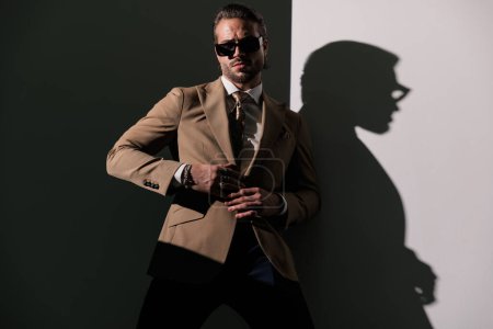 Photo for Elegant young man with sunglasses adjusting brown suit and posing while looking forward in front of grey background - Royalty Free Image