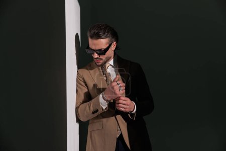 Foto de Retrato de hombre de moda elegante en traje marrón mirando hacia abajo y ajustando pulseras mientras se acuesta y se coloca en una pared sobre fondo gris - Imagen libre de derechos