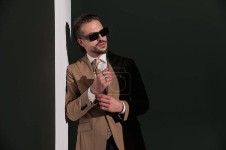 Foto de Hombre fresco sin afeitar en traje marrón la fijación de pulseras y mirando a un lado, mientras que la colocación en una pared - Imagen libre de derechos