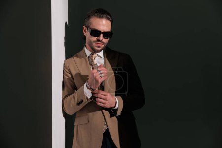 Foto de Hombre confiado en traje marrón ajustando pulseras y acostado en una pared gris en el estudio - Imagen libre de derechos